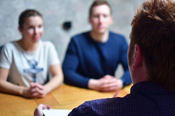 Zwei Personen sitzen an einem Tisch und hören einem Gesprächspartner zu.