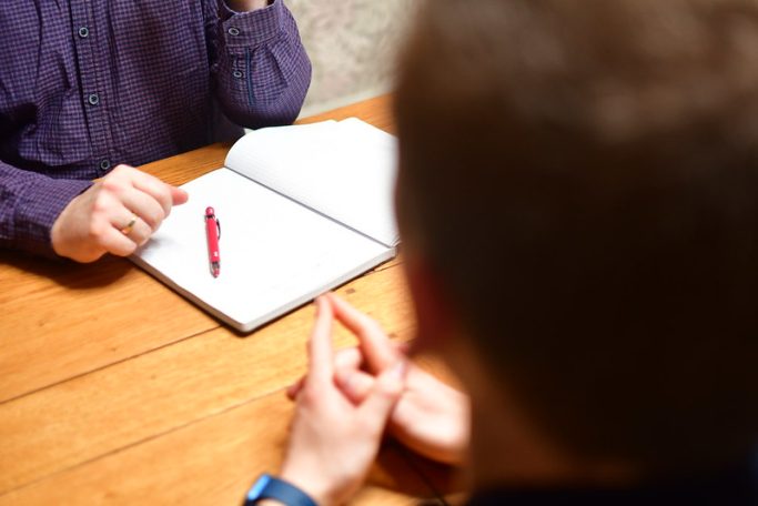 Zwei Personen sitzen an einem Tisch, eine hält einen Stift, eine Notizbuch liegt offen.